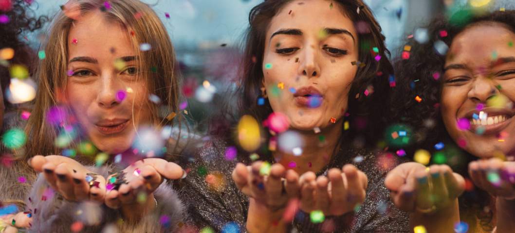 Young women blowing confetti from hands. Friends celebrating outdoors in evening at a terrace.