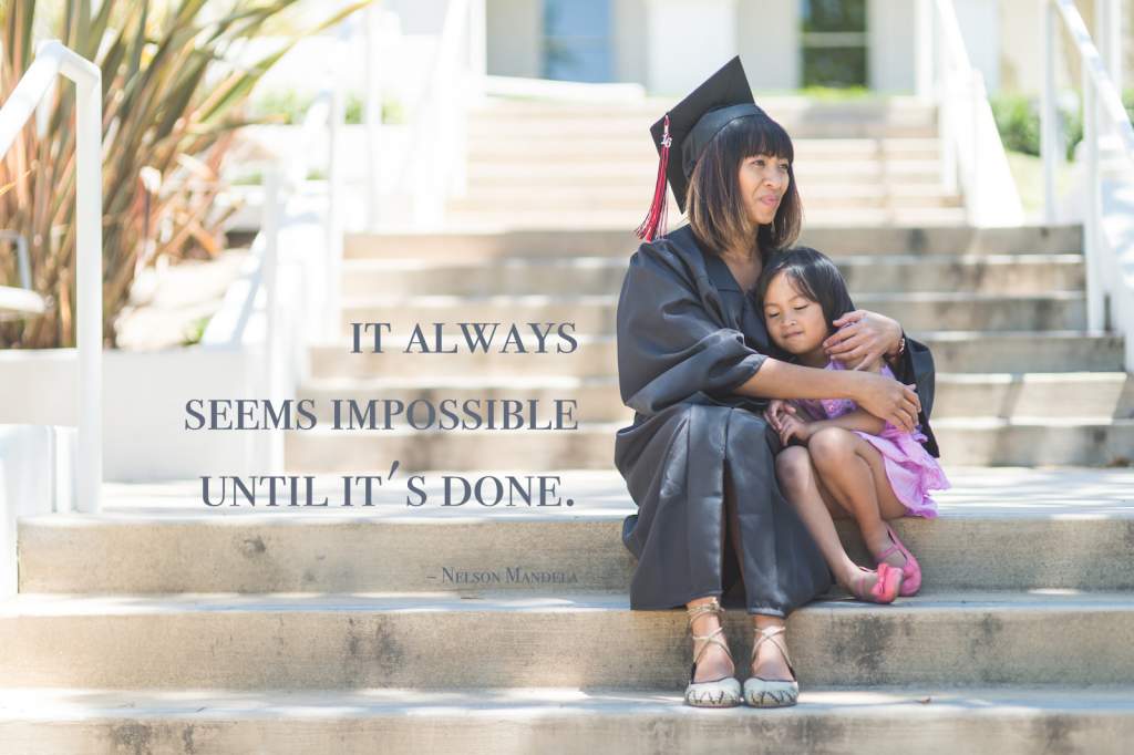 woman in graduation robe and cap sits on steps with her young daughter after graduation. 