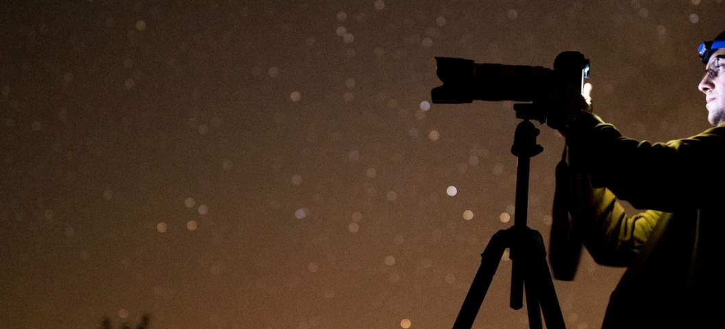 Astral photography for beginners is demonstrated by a man, his camera, and tripod in the night sky