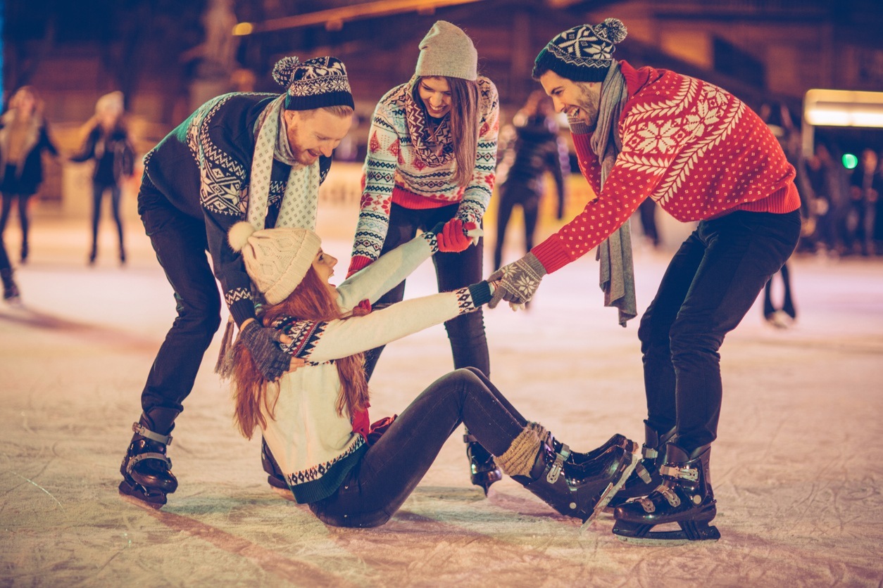 Friends having so much fun while ice skating. Wearing warm clothing. City is decorated with christmas lights.
