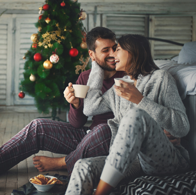 couple laughing and drinking coffee in front of a christmas tree.