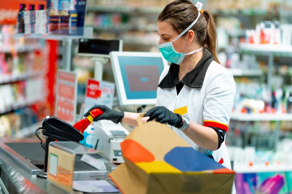 Woman cashier wearing protective face mask and gloves to prevent viruses, scanning disinfection products at the cash register and packing in paper bag