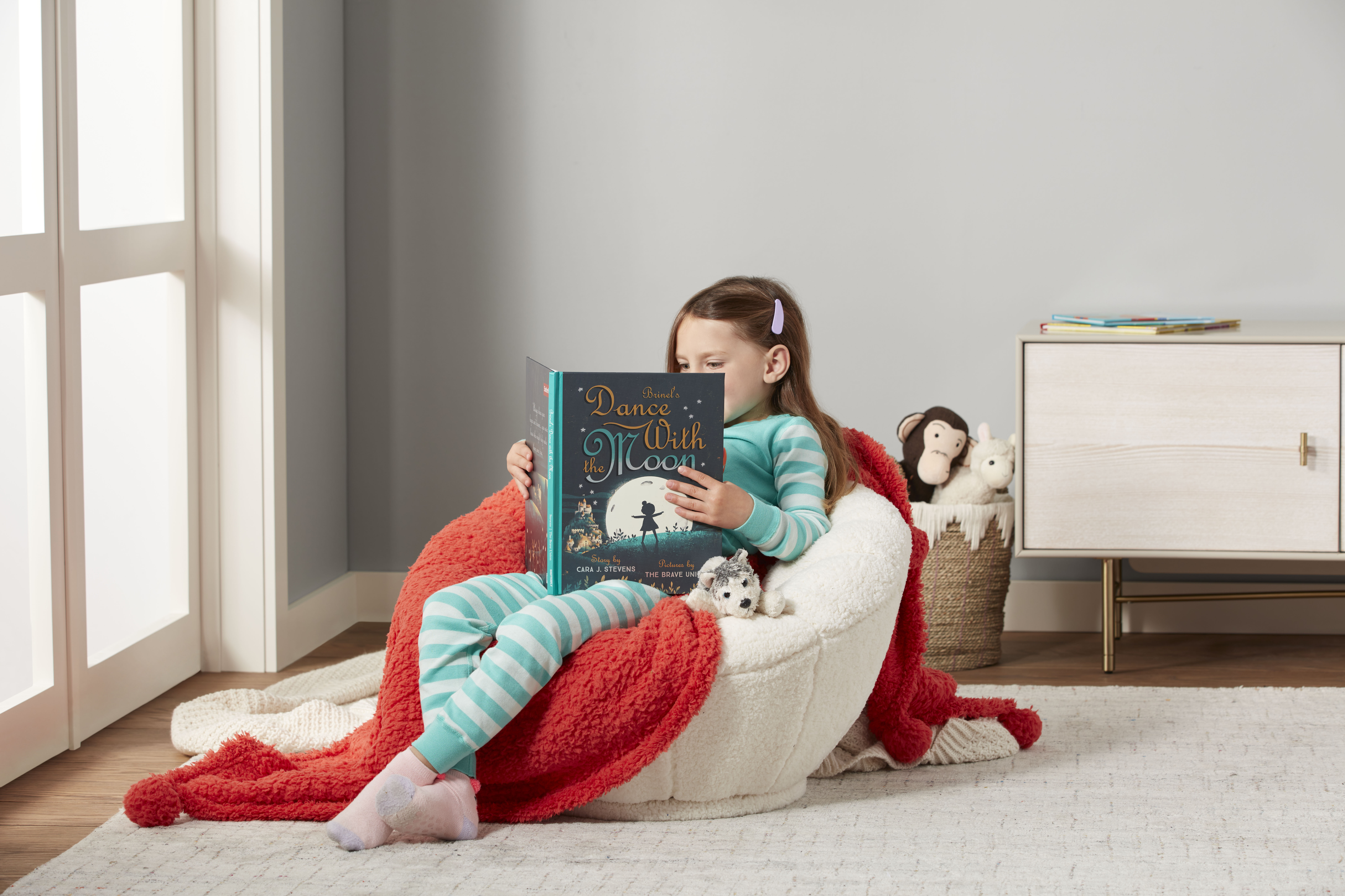 Little girl reading a story book on a beige personalized bean bag chair featuring a red blanket and a basket of stuffed animals