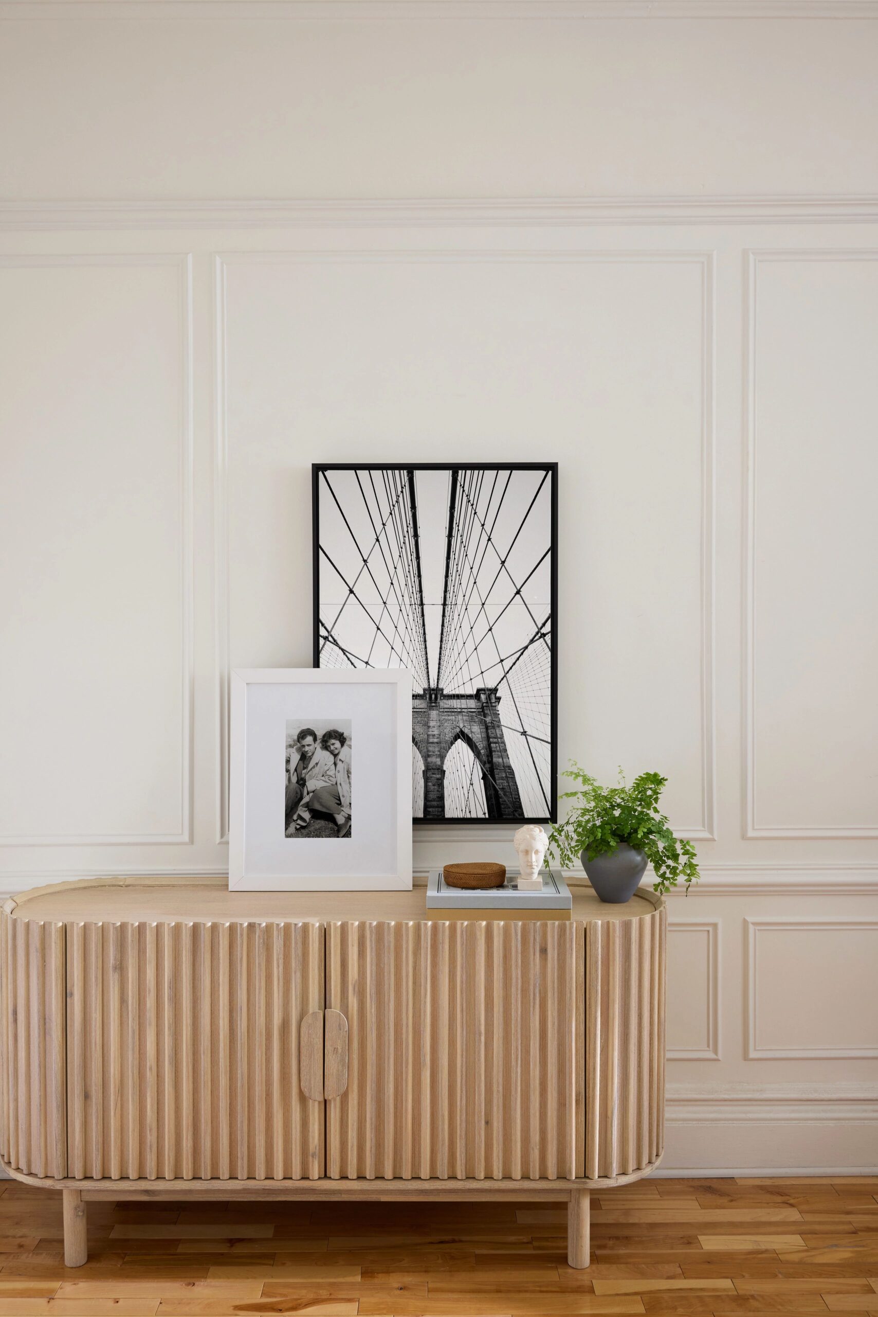 A minimalist interior features a wooden sideboard adorned with a framed canvas print with white mat, a black framed print, a potted plant, standing gold floor lamp, white lampshade, and decorative objects, set against a white paneled wall.